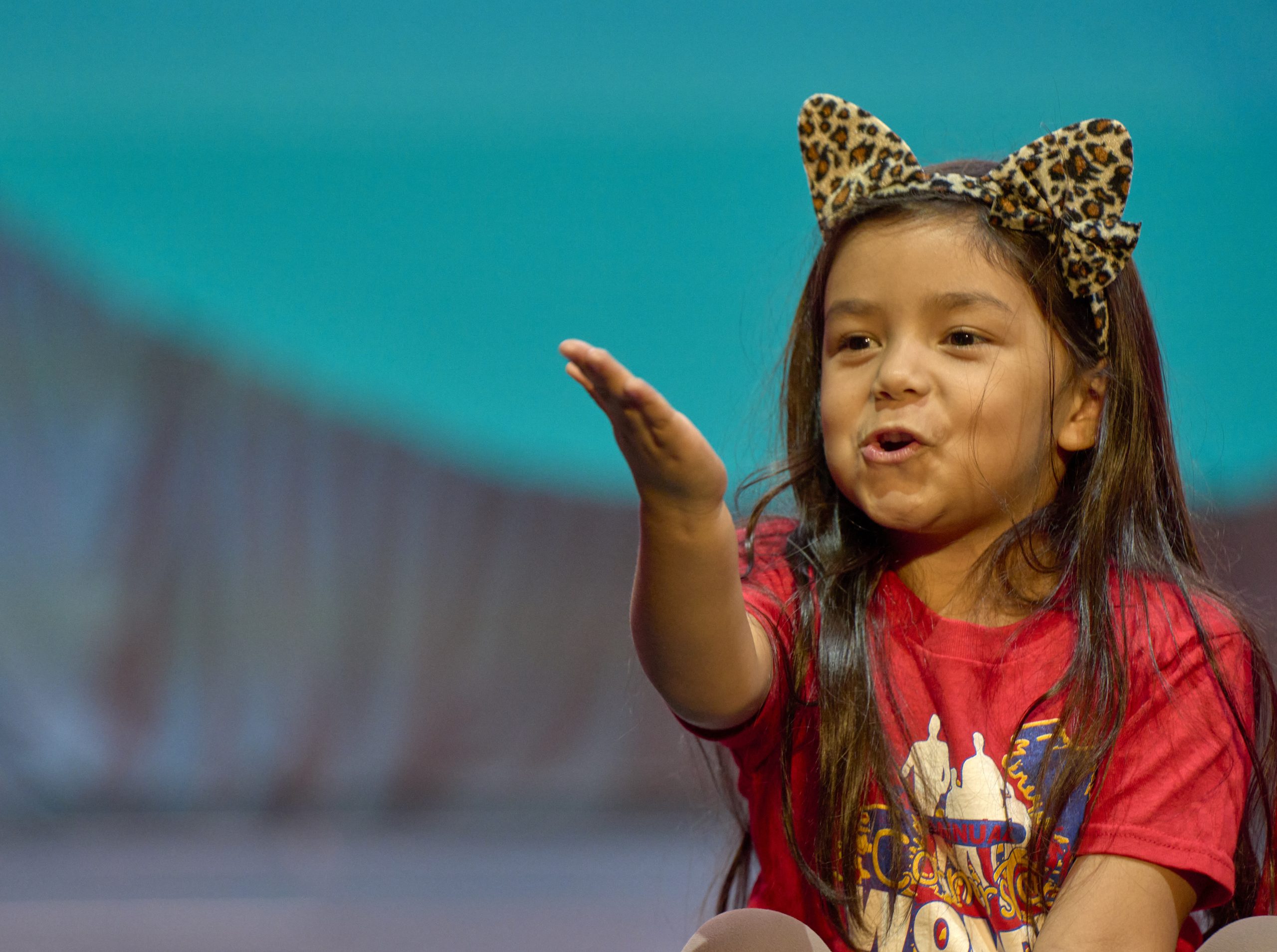 A young girl in a red shirt and cat ear headband blows a kiss from a stage