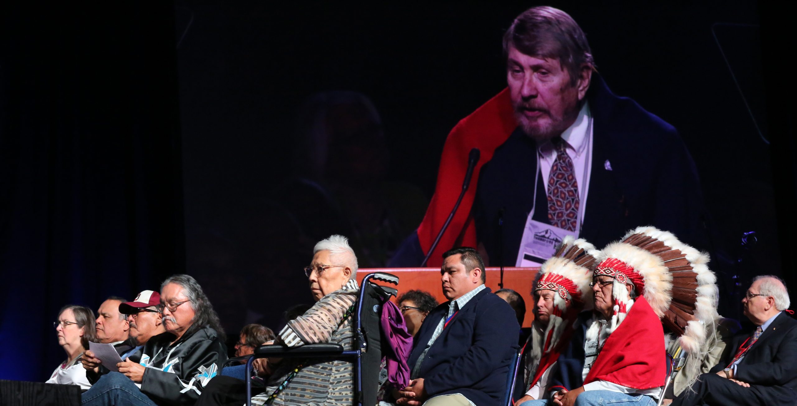 Arapaho and Cheyenne Tribal Representatives in traditional dress sit on a stage along with others involved in the Sand Creek Massacre Report listen to Gary L. Roberts (on screen behind those seated) at the 2016 United Methodist General Conference in Portland, Ore. Photo by Kathleen Barry, UMNS