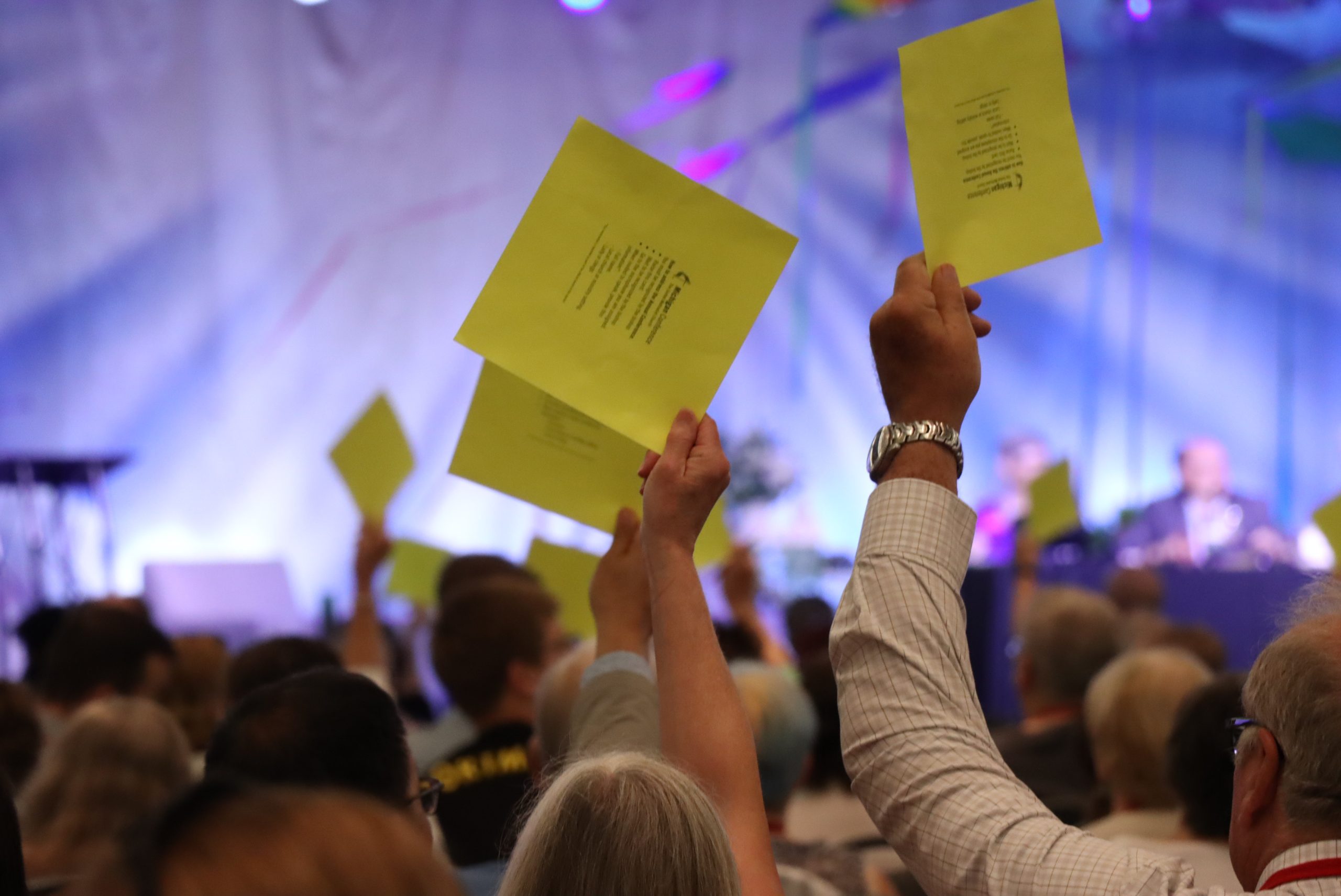 People in a crowd hold up yellow pieces of paper against a blue and white backdrop. The individuals are raising the papers to vote or be called on during the 2024 business meeting of the Michigan Conference of The United Methodist Church.