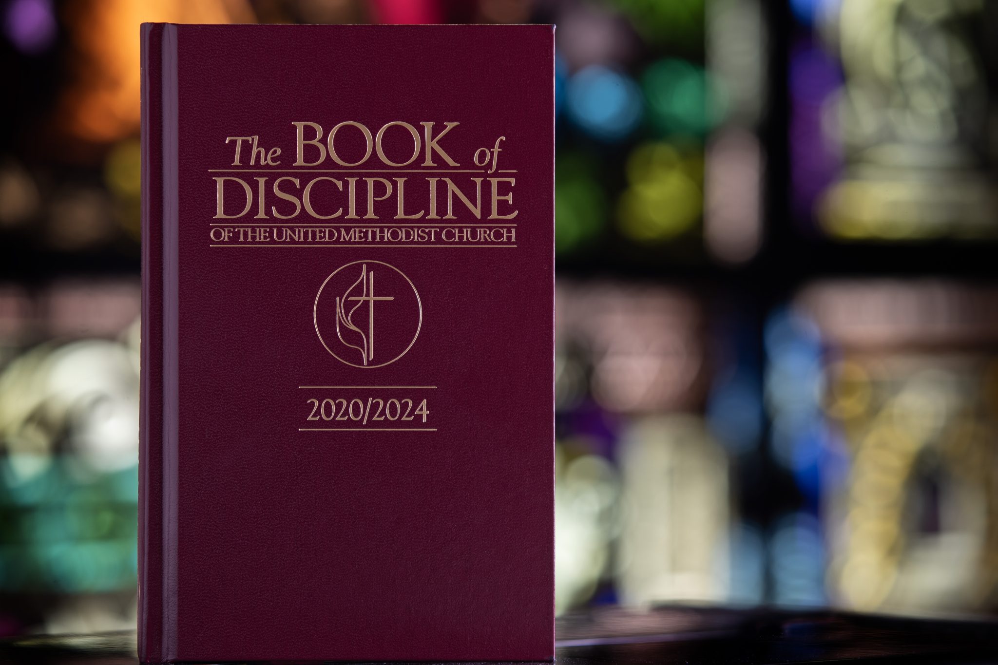 The maroon Book of Discipline of The United Methodist Church (2020/2024) stands upright in focus in front of a faded background of stained glass windows