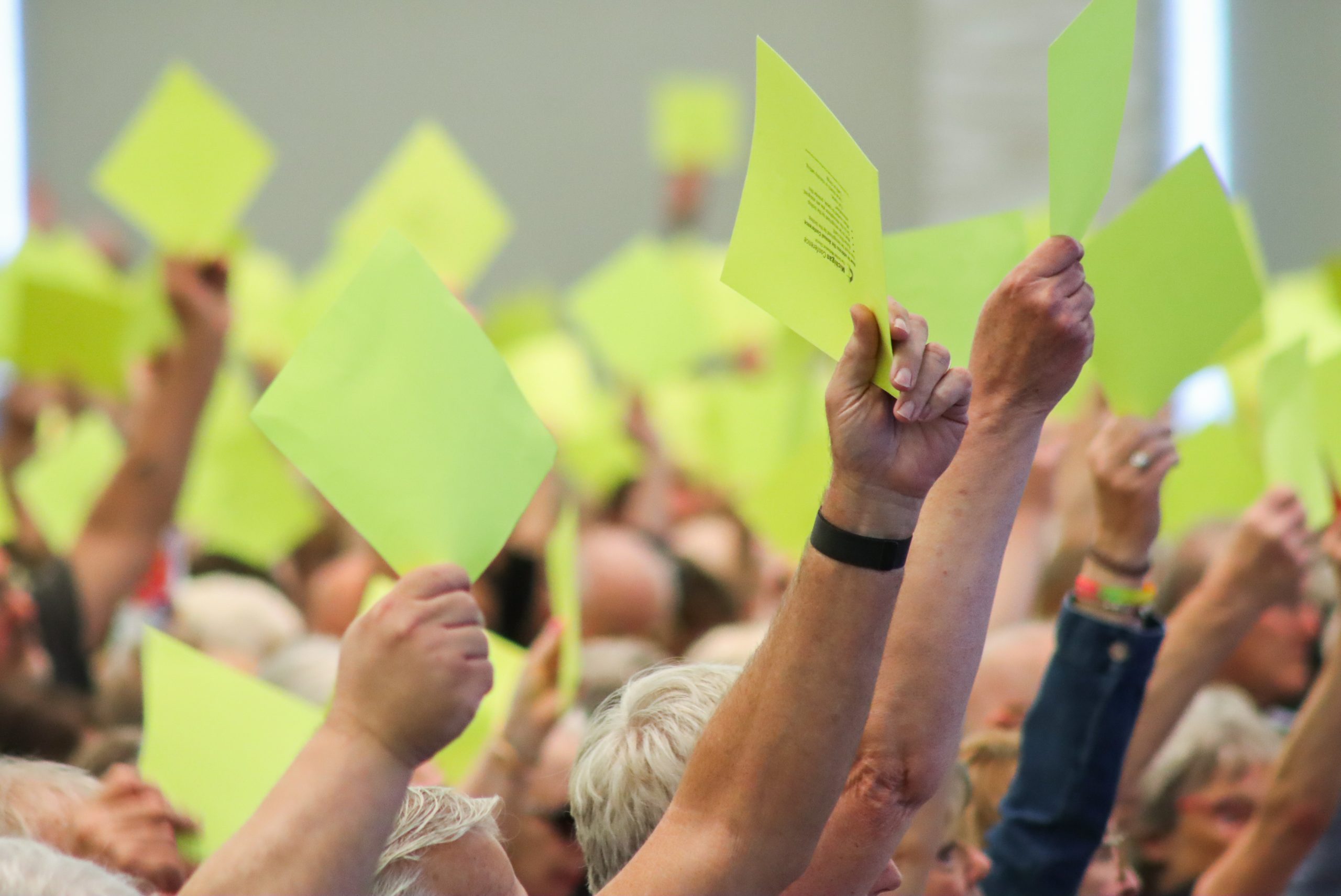 Many arms are raised holding bright yellow pieces of paper as they vote at a conference